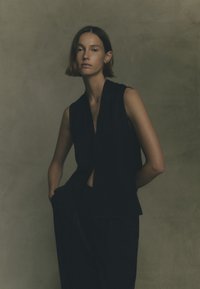 Woman with short brown hair wearing a sleeveless black vest and pants, standing with hands in pockets against a plain dark background.