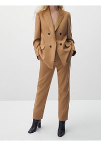 En person står självsäkert i en stilren, kroppsnära beige kostym med svarta knappar och höga svarta stövlar, mot en minimalistisk bakgrund.