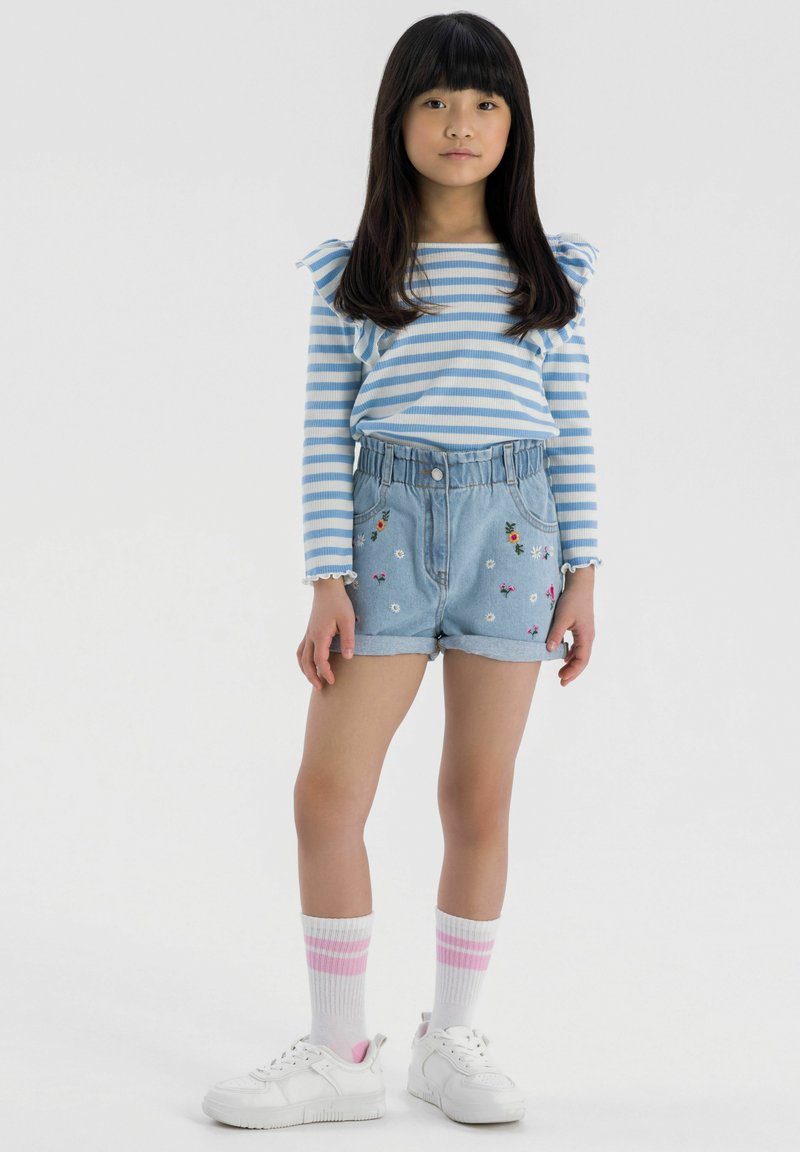 Blau-weiß gestreiftes Langarmhemd mit Volants an den Schultern, kombiniert mit hellblauen Jeansshorts mit floralem Stickereien und einem gerafften Bund.