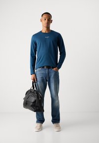 Jonge man in een blauwe blouse met lange mouwen, jeans en beige sneakers, met in een hand een zwarte leren tas en de andere hand in zijn zak.