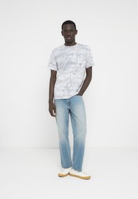 Γκρι γραφικό t-shirt με αφηρημένο σχέδιο, κοντά μανίκια, συνδυασμένο με ανοιχτό μπλε τζιν παντελόνι και λευκά αθλητικά παπούτσια με καφέ σόλες.