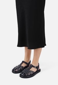 Sandalias de cuero negro con un diseño recortado, adornadas con tachuelas plateadas, que cuentan con una correa en el tobillo y una suela plana.