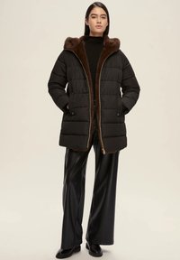 Cappotto piumino nero con cappuccio foderato in pelo marrone, chiusura con zip e motivo trapuntato, abbinato a pantaloni neri a gamba larga lucidi e stivali.