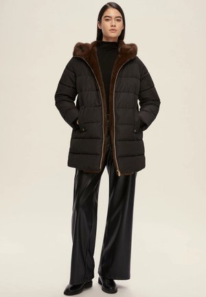 Cappotto piumino nero con cappuccio foderato in pelo marrone, chiusura con zip e motivo trapuntato, abbinato a pantaloni neri a gamba larga lucidi e stivali.