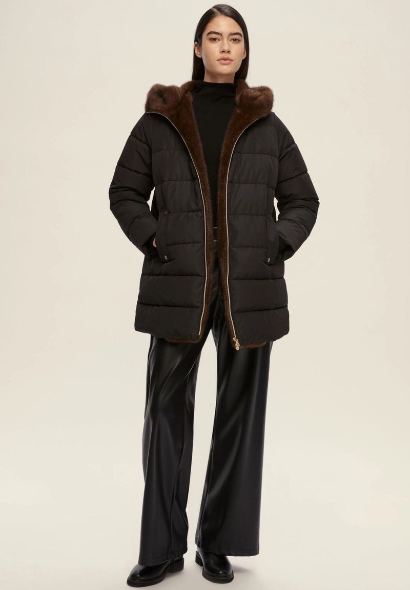 Cappotto piumino nero con cappuccio foderato in pelo marrone, chiusura con zip e motivo trapuntato, abbinato a pantaloni neri a gamba larga lucidi e stivali.