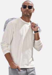 Langärmliges cremefarbenes Hemd aus leichtem Stoff, kombiniert mit grauen Hosen. Die Accessoires umfassen schwarze Sonnenbrillen und einen hellgrauen Rucksack.