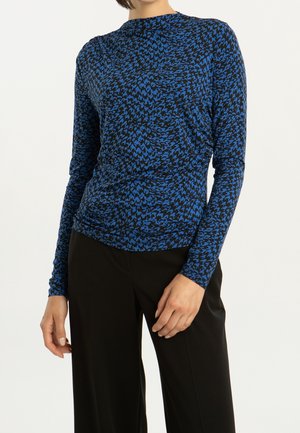 Kobieta w niebiesko-czarnej bluzce z abstrakcyjnym wzorem na długim rękawie i prostych czarnych spodniach, pozuje z jedną ręką przy udzie.