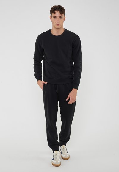 Felpa nera e joggers realizzati in morbido misto cotone, con polsini a coste e dettaglio del logo sulla manica. Abbinati a sneakers bianche.