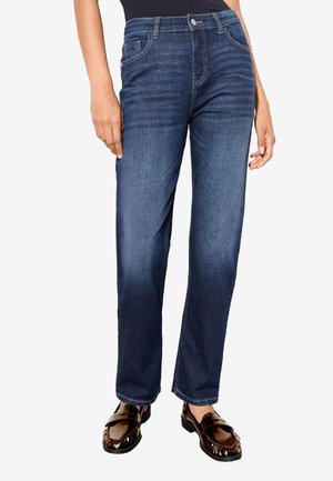 Žena nosiace modré rifle s rovnými nohavicami a tmavohnedé lesklé mokasíny, stojaca s uvoľnenými rukami pozdĺž tela.