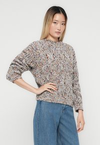 Jonge vrouw met lang ombre-haar, gekleed in een grijze gebreide trui met kleurrijke spikkels en een paar blauwe wijde jeans, poseert met één hand op de heup.