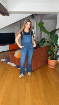 Νεαρή γυναίκα που χαμογελά με τα χέρια στις τσέπες, φοράει τζιν σαλοπέτα και μαύρο πουκάμισο, στέκεται σε σαλόνι με καφέ καναπέ και μεγάλο φυτεμένο φυτό.