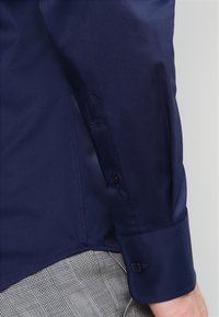 Dunkelblaues Hemd mit glatter Textur, Knopfmannschäften und präzisen Nähten. Verfügt über eine taillierte Passform und elegante Designelemente.