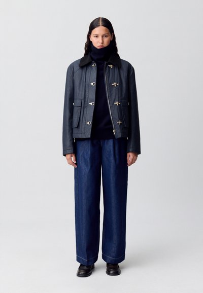 Veste en cuir bleu marine avec des accessoires en or, dotée d'une fermeture éclair et de boutons à bascule, associée à un pantalon en denim ample et des chaussures noires.