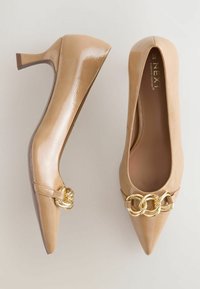 Beige patent leather korkokengät, joissa on terävä kärki ja kultaketjun yksityiskohtia, esiteltynä vaaleaa taustaa vasten.