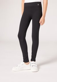 Leggings negros de canalé con una cinturilla ancha, combinados con zapatillas blancas con tiras de velcro y suelas texturizadas.