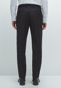 Donkergrijze op maat gemaakte broek met een slim fit, een gladde textuur, twee achterzakken en een subtiele plooival die langs de voorkant loopt. Zwarte geklede schoenen.