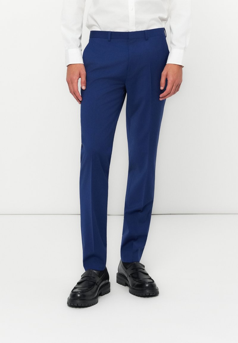 Pantalones azul ajustados, hechos de una tela suave, con frente plano y dos bolsillos laterales. Se llevan con zapatos negros y una camisa blanca.