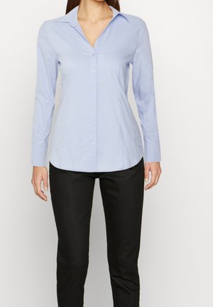 Woman wearing light blue buttoned long-sleeve shirt with collar, paired with black pants, standing against a plain white background.