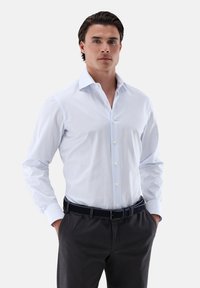 Camicia button-up a righe blu chiaro con colletto, maniche lunghe e bottoni bianchi, abbinata a pantaloni scuri e una cintura coordinata.