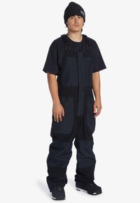 Zwarte snowboardoverall met verstelbare banden, meerdere zakken, een contrasterende stoftextuur en een losse pasvorm. Gedragen met een zwarte beanie.
