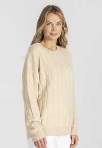 Beżowy sweter z dzianiny z wzorem warkocza, okrągłym dekoltem i ściągaczami przy rękawach. Materiał wydaje się miękki i teksturowany, oferując luźny krój.