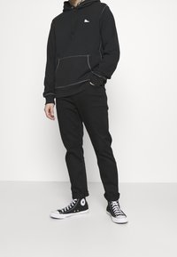 Felpa nera con cuciture bianche e logo, tasca a marsupio, abbinata a pantaloni neri e sneakers alte nere.