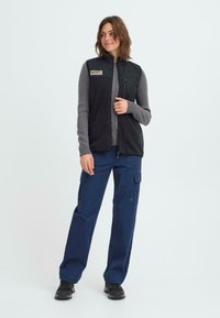 Schwarze Fleece-Weste über einem grauen Pullover, kombiniert mit lockeren, navyblauen Cargo-Hosen. Die Weste hat einen Reißverschluss vorne und Seitentaschen. Schwarze Schuhe runden das Outfit ab.