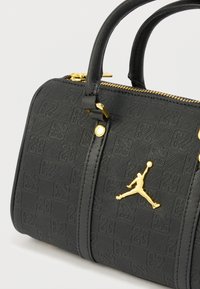 Musta käsilaukku, jossa kohokuvioitu pinta, kullanväriset metalliosat, kaksinkertaiset kahvat ja vetoketjusuljenta. Koristeena kullanvärinen Air Jordan -embleemi.
