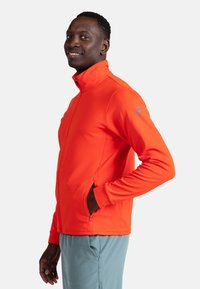 Lys orange zip-jakke med høj krave, strækbart stof, sidelommer og et logo på venstre skulder. Bæret over lyseblå bukser.
