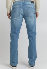 Jeans de denim azul claro con un corte recto, que cuentan con dos bolsillos traseros y una sutil textura deslavada. Zapatillas blancas visibles en el dobladillo.