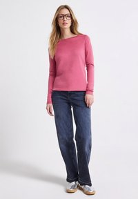 Różowa bluzka z długim rękawem i okrągłym dekoltem, zestawiona z ciemnoniebieskimi, luźnymi dżinsami oraz srebrnymi trampkami. Włosy rozpuszczone, nosi duże okulary.