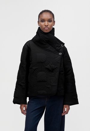 Femme portant une veste matelassée noire avec un col haut asymétrique et un jean bleu foncé sur un fond clair uni.
