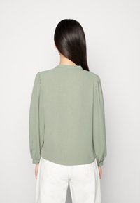 Blusa verde chiaro con maniche lunghe a sbuffo e colletto rotondo. Texture liscia con sottili plissettature sulle cuciture delle spalle. L'orlo è leggermente curvato.