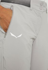 Pantalons gris clair en matériau doux, dotés d'une poche zippée, d'une taille à motif noir et d'un logo blanc près de la poche.