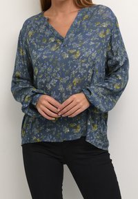 Blouse florale bleue avec décolleté en V, manches longues et coupe ample. Présente un tissu texturé avec des motifs floraux verts et violets.