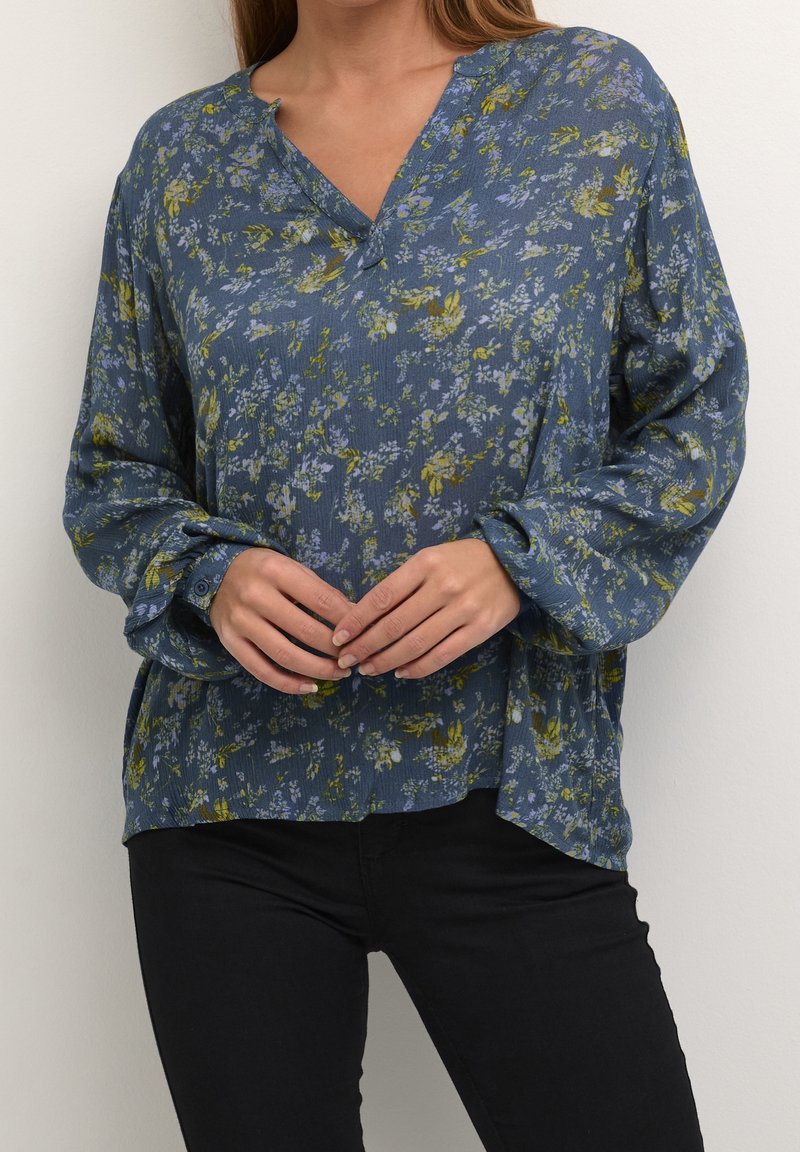 Blouse florale bleue avec décolleté en V, manches longues et coupe ample. Présente un tissu texturé avec des motifs floraux verts et violets.