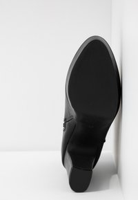 Black high-heeled shoe with a pointed toe, featuring a smooth leather upper and a textured rubber sole, angled against a white background.