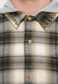 Καρό φανελένιο πουκάμισο με γκρι κουκούλα, διακοσμημένο με χρυσά τρουκ στο γιακά, σε μπεζ και πράσινους τόνους, με ξύλινα κουμπιά. Υφή υφάσματος με ορατά ραφές.