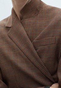 Bruine gewatteerde wikkeljas met een hoge kraag, gemaakt van een gestructureerde stof. Het ontwerp bevat een houndstooth-patroon in verschillende tinten.
