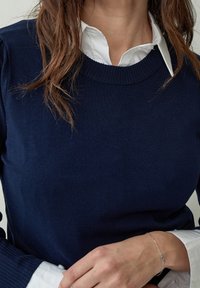 Pull en maille bleu marine avec un col côtelé, porté par-dessus une chemise blanche à col. Gros plan sur le haut du corps et une partie du bras, mettant en valeur la texture.