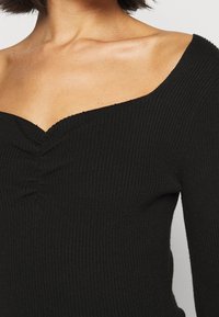 Kvinna klädd i en svart ribbad off-shoulder-topp med drapering vid den mellersta halsringningen.