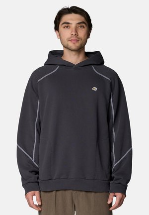 Junger Mann trägt ein dunkelgraues Kapuzensweatshirt mit kontrastreichen Nähten und einem kleinen Logo auf der linken Brustseite.