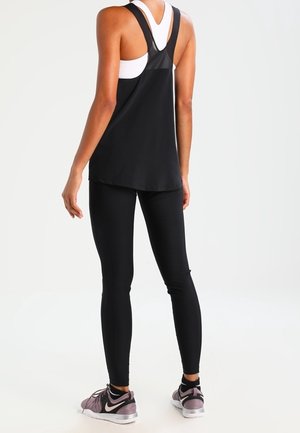 Kvinde iført sort tanktop og leggings med lyserøde og sorte sportssko, stående med ryggen til på ensfarvet baggrund.