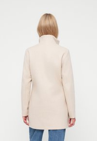 Cappotto di lana beige con colletto alto e vestibilità su misura, caratterizzato da maniche lunghe e una texture morbida. Indossato sopra jeans blu.
