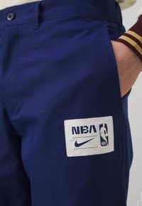 Marineblå stoffshorts med en glatt tekstur, utstyrt med en hvit NBA-logoapplikasjon og et Nike-logo. Designet med knapp i midjen og sidelommer.