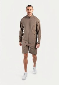 Beige atletisch jack met volledige ritssluiting en hoge kraag, gecombineerd met bijpassende korte broek. Model draagt witte sneakers. Glad, lichtgewicht materiaal.