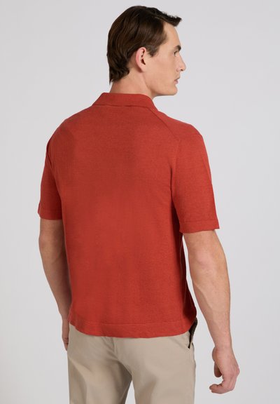 Homme de dos portant un polo à manches courtes couleur rouille et un pantalon beige sur un fond uni gris clair.
