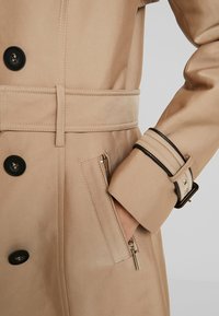 Beżowy trencz z czarnymi guzikami, teksturowanej tkaniny, z paskiem w talii i zamykaną kieszenią na zamek. Posiada ozdobne mankiety z detalami.