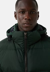 Dark green puffer jacket with a high collar, front zipper, and adjustable hood. Smooth texture, featuring black hardware and branding detail.