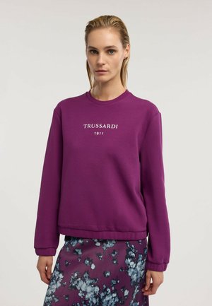 Frau trägt einen lila Pullover mit dem Text "Trussardi 1911" und einen lila Rock mit blau-weißen Blumenmustern, steht vor einem einfarbigen Hintergrund.